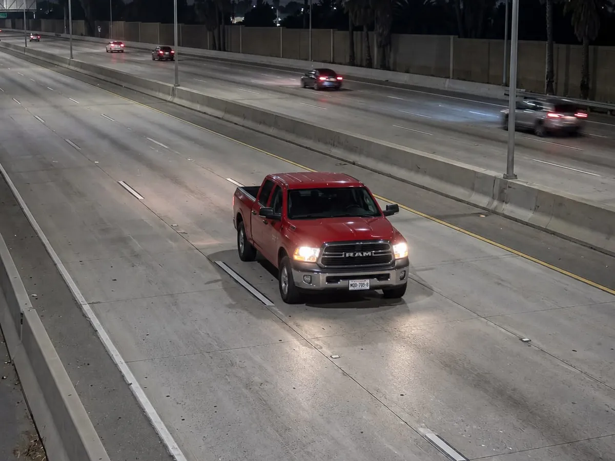 A red crew-cab pickup truck drives toward the camera with its headlights on, in the slow lane of a lit San Diego freeway at night, seen from a fixed ALPR camera on a mast arm above the shoulder, its front plate legible under a light pool.